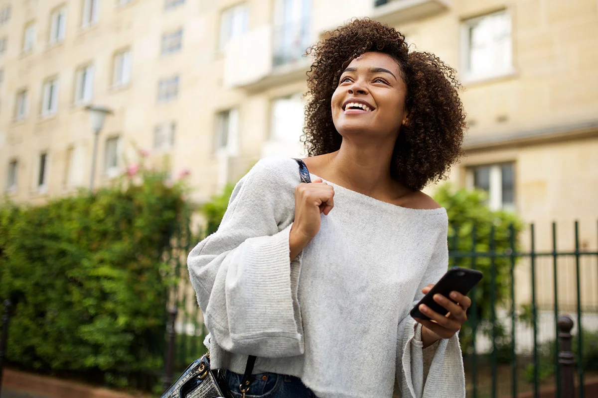 smiling woman holds her phone with one hand and her bag strap with the other while searching for addiction resources