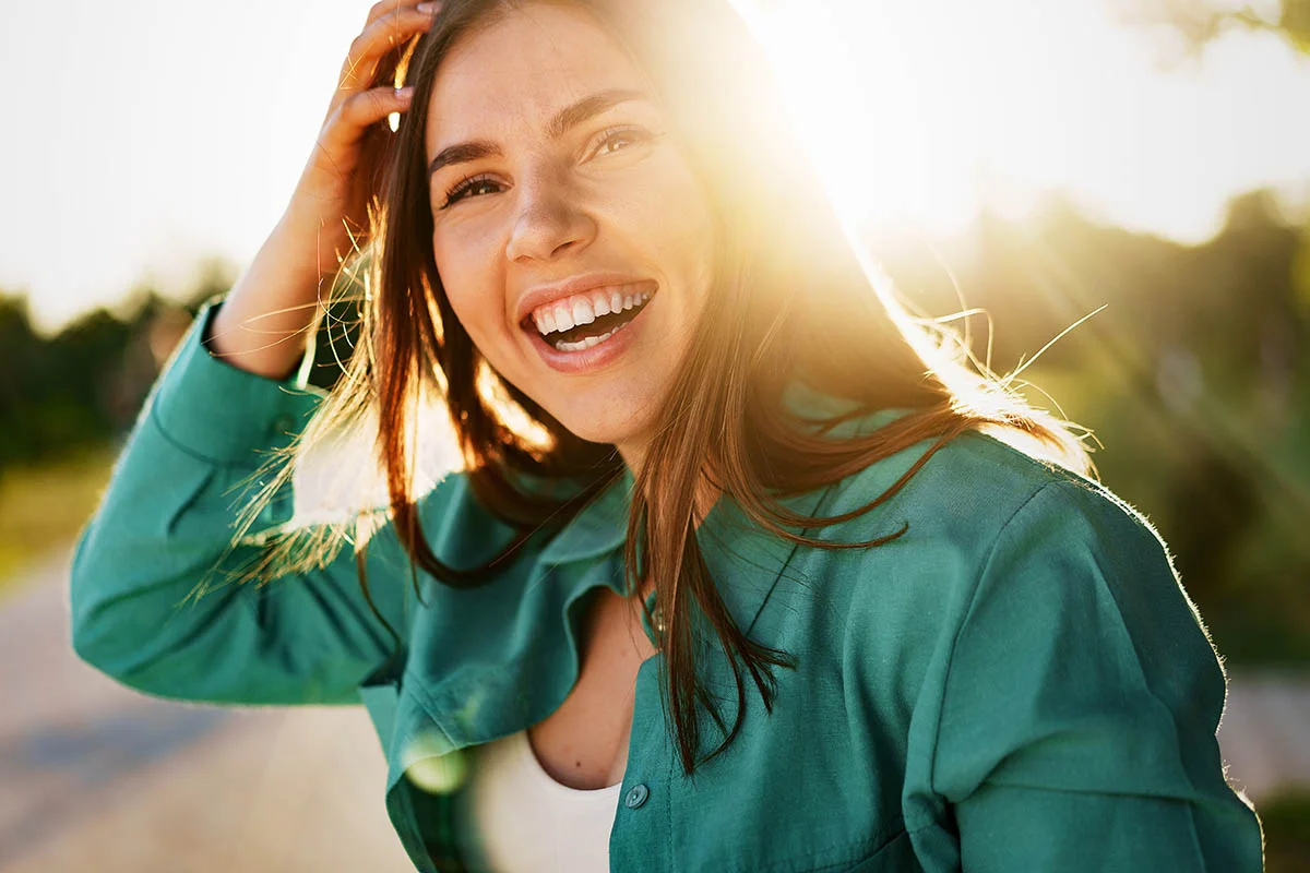 smiling woman stands outsides during sunset and touches her hand to the top of her head
