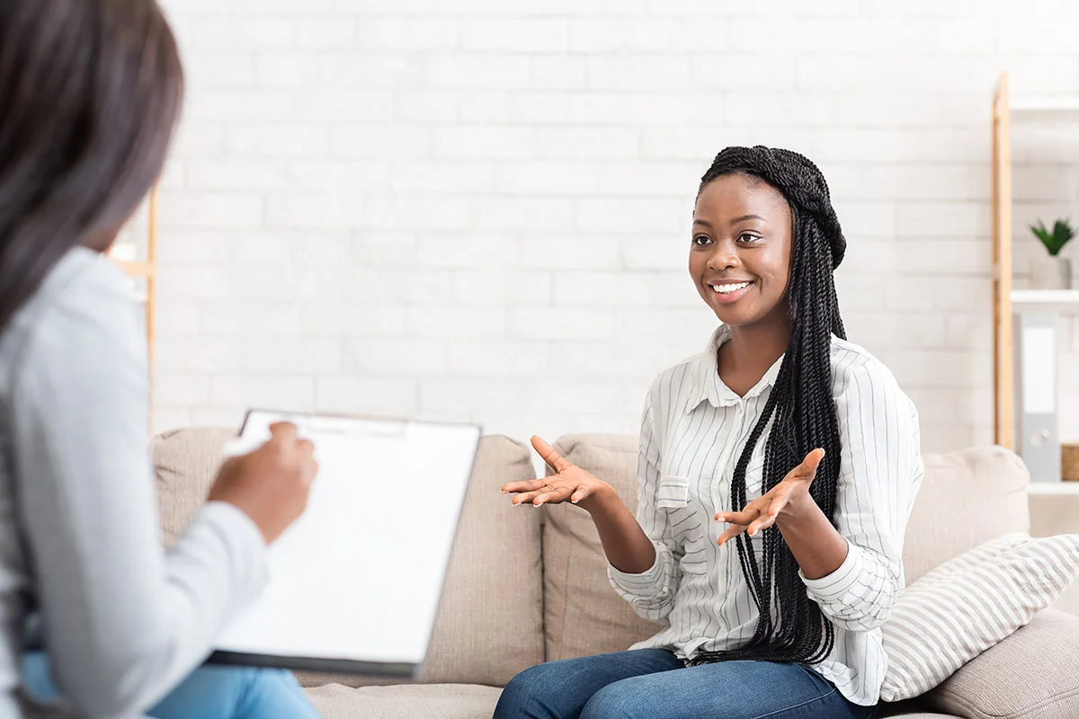 smiling woman sits on a couch while talking to her therapist who is taking notes